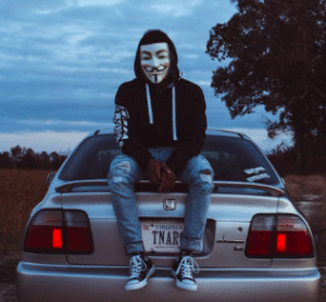Hacker in mask sitting on car, representing carmaker portal security flaw
