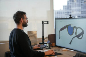 A designer works on a CAD model of the Sesame conversational AI smart glasses on a large monitor.